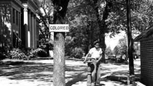 A black boy using a water fountain beneath a "colored" sign during the Jim Crow era