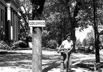 A black boy using a water fountain beneath a "colored" sign during the Jim Crow era